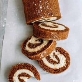 A rolled cake with a brown exterior and white creamy filling, sliced into several pieces, placed on parchment paper with a dusting of powdered sugar.