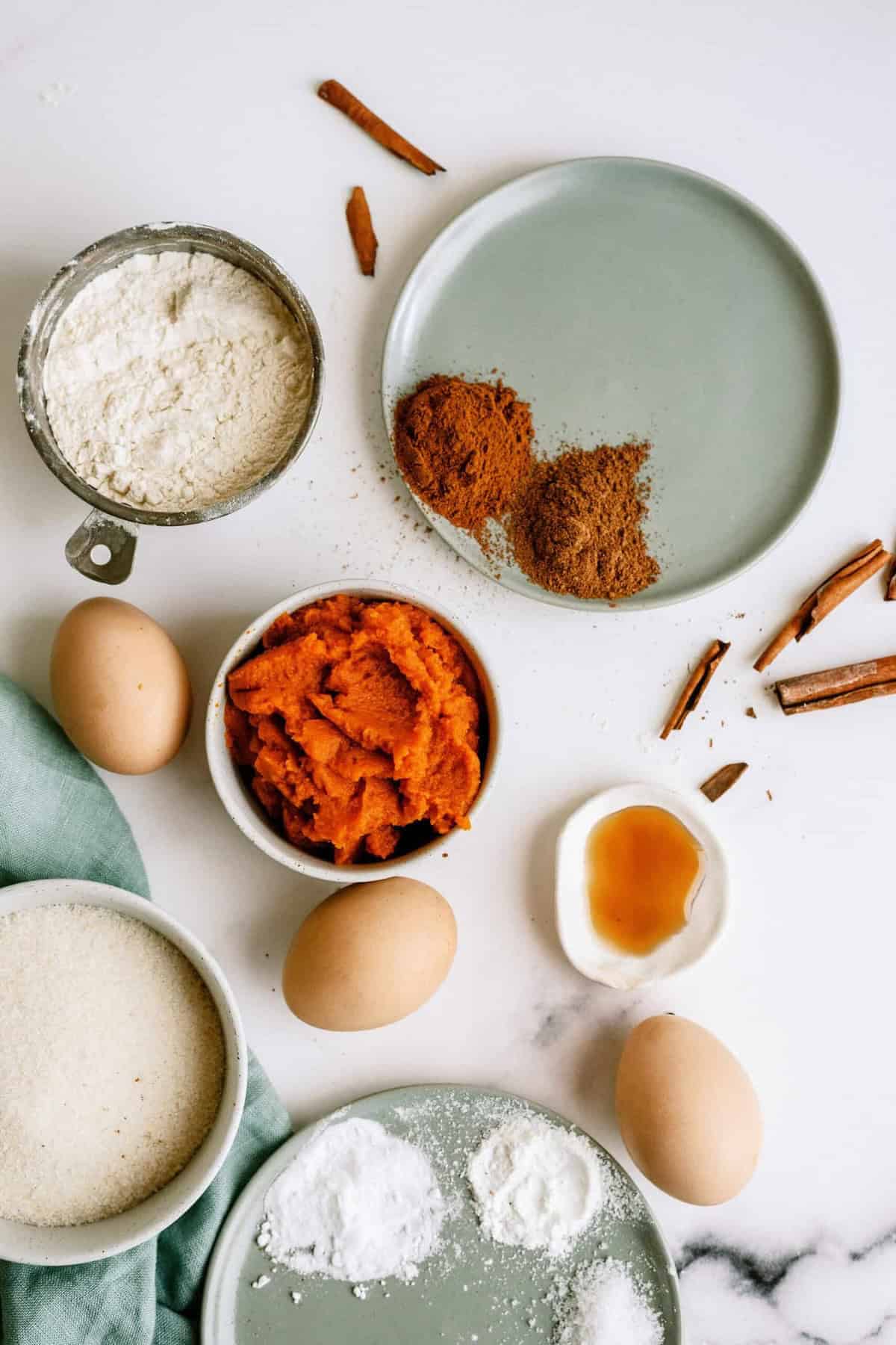 Ingredients for baking are laid out on a white surface, including flour, sugar, eggs, pumpkin puree, vanilla extract, baking powder, baking soda, and ground spices.