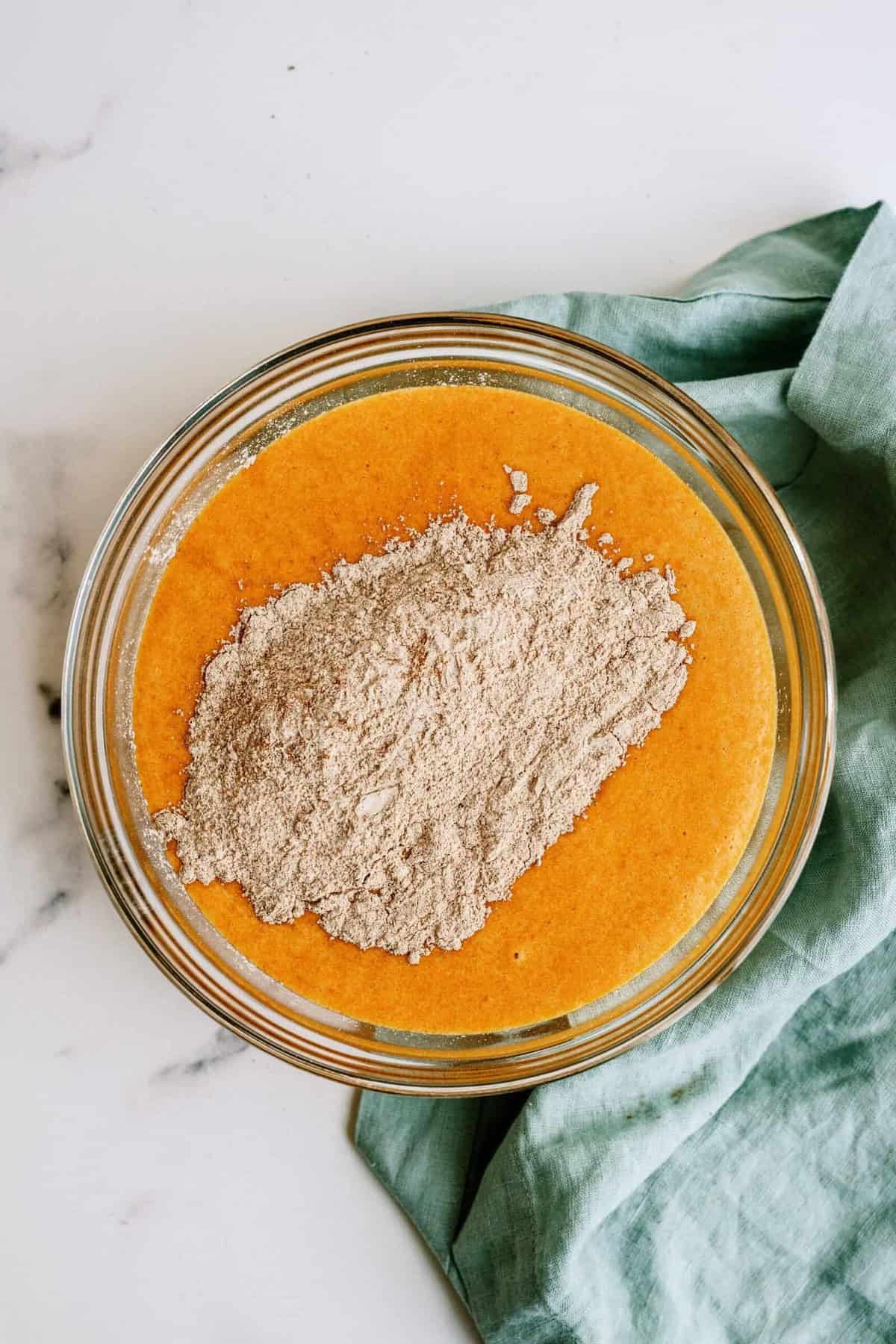 A glass bowl containing an orange-colored liquid mixture with a pile of flour on top, placed on a green cloth on a white surface.