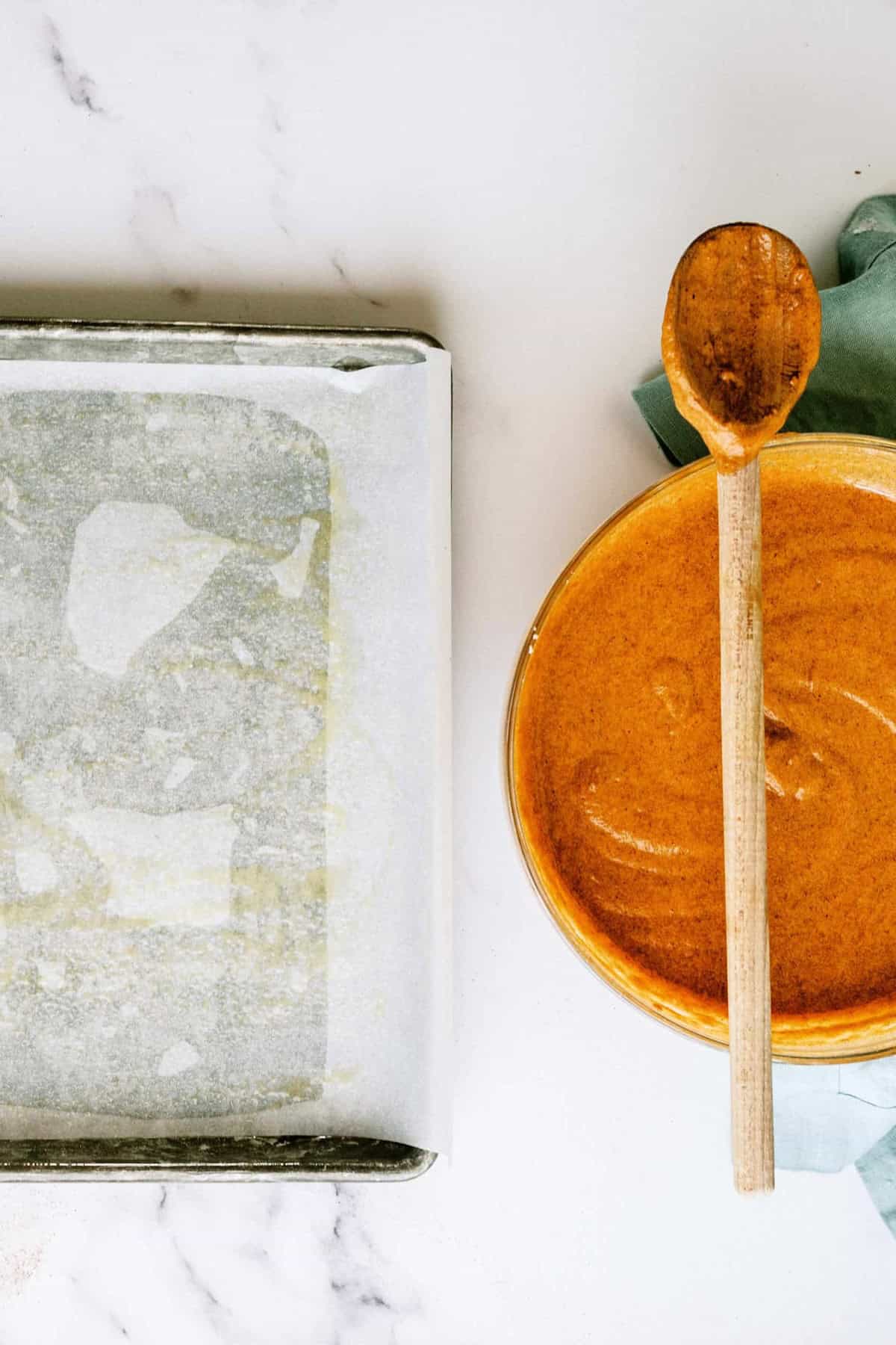 A baking sheet with parchment paper on the left and a bowl of batter with a wooden spoon resting on it on the right.