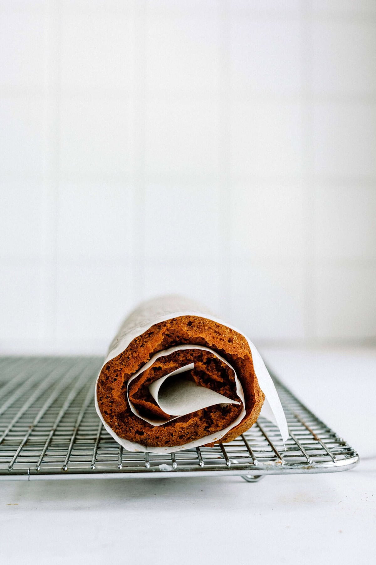 A rolled cake with parchment paper wrapped around it sits on a wire cooling rack against a tiled white background.