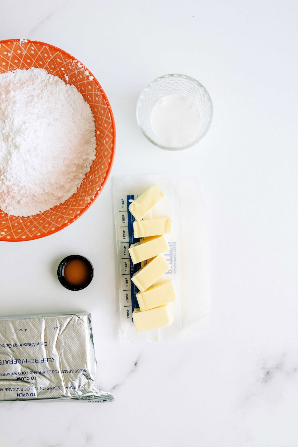 Overhead view of baking ingredients: a bowl of powdered sugar, a glass of liquid, a capful of vanilla extract, four sticks of butter, and an opened foil-wrapped butter package on a white surface.