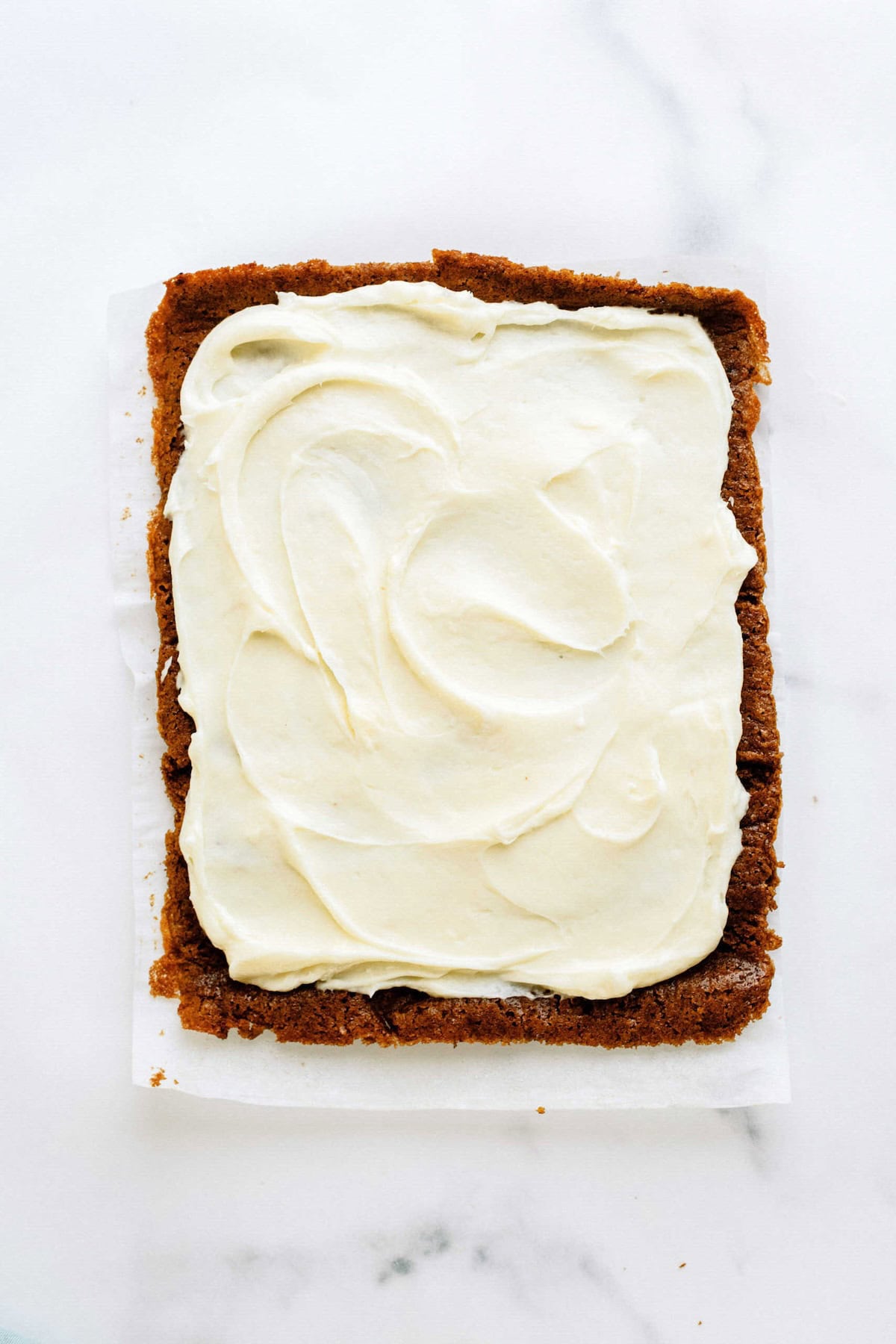 A rectangular piece of cake on parchment paper, topped with a thick layer of white frosting, placed on a white surface.