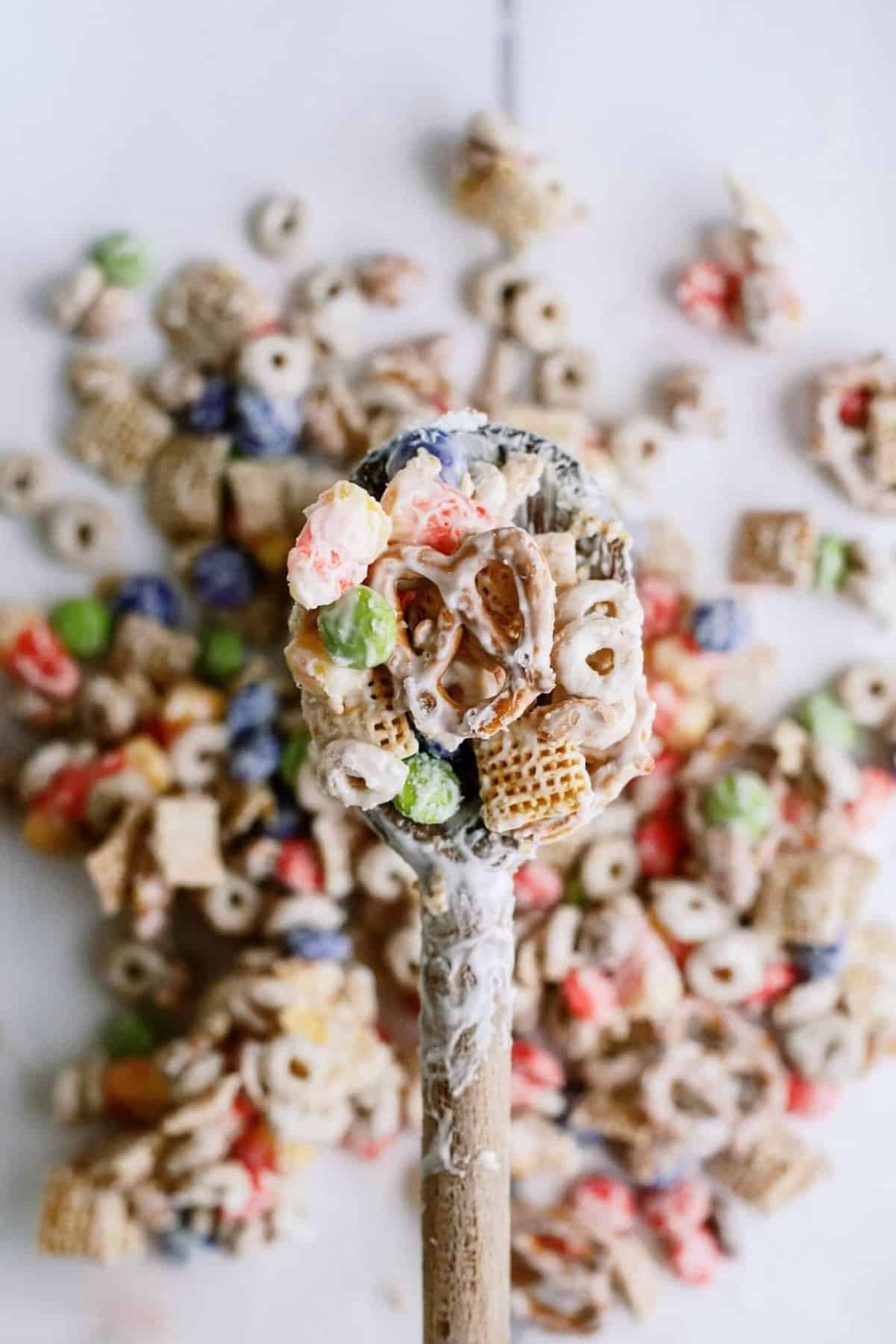 A wooden spoon holds a mix of colorful cereal pieces coated in a white candy-like coating, with more cereal scattered in the background.