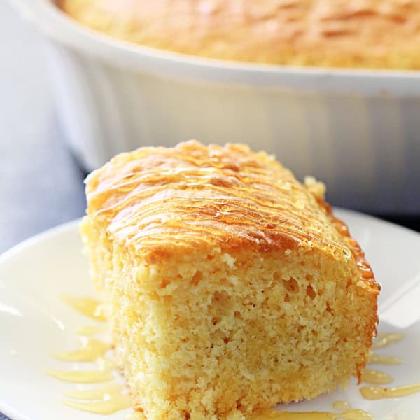 A slice of cornbread on a white plate with the remaining cornbread in a baking dish in the background.