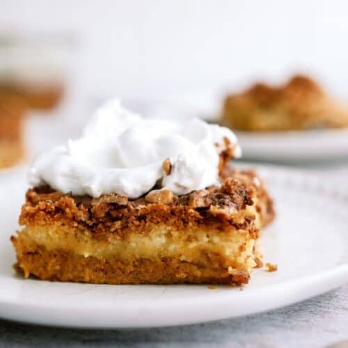 A slice of dessert with a crumbly top and a layer of cream, topped with whipped cream, sits on a white plate.