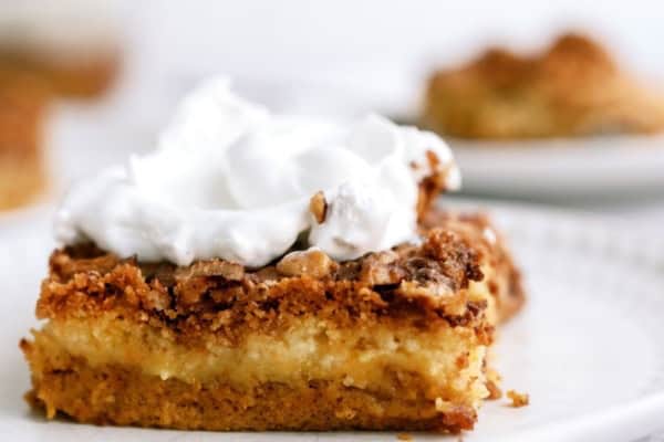 A slice of dessert with a crumbly top and a layer of cream, topped with whipped cream, sits on a white plate.