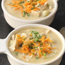 Two white bowls of creamy turkey soup topped with shredded cheese and chopped chives on a dark surface.