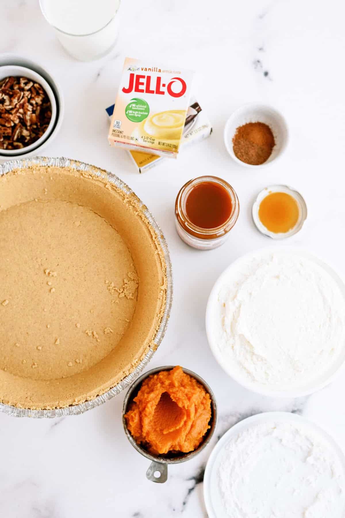 Ingredients for a pumpkin dessert including a pie crust, JELL-O vanilla pudding mix, canned pumpkin, pecans, spices, vanilla extract, caramel sauce, and bowls of flour and sugar arranged on a white surface.