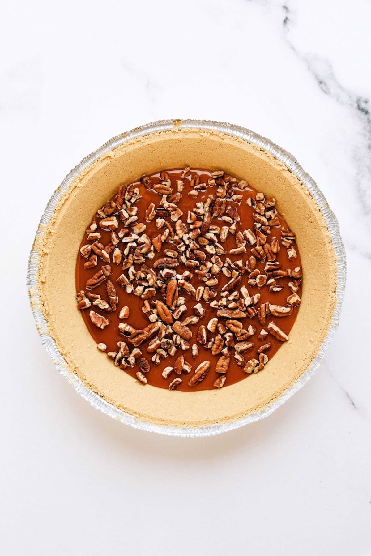 An unbaked pie crust filled with caramel and topped with chopped pecans sits on a white marble surface.