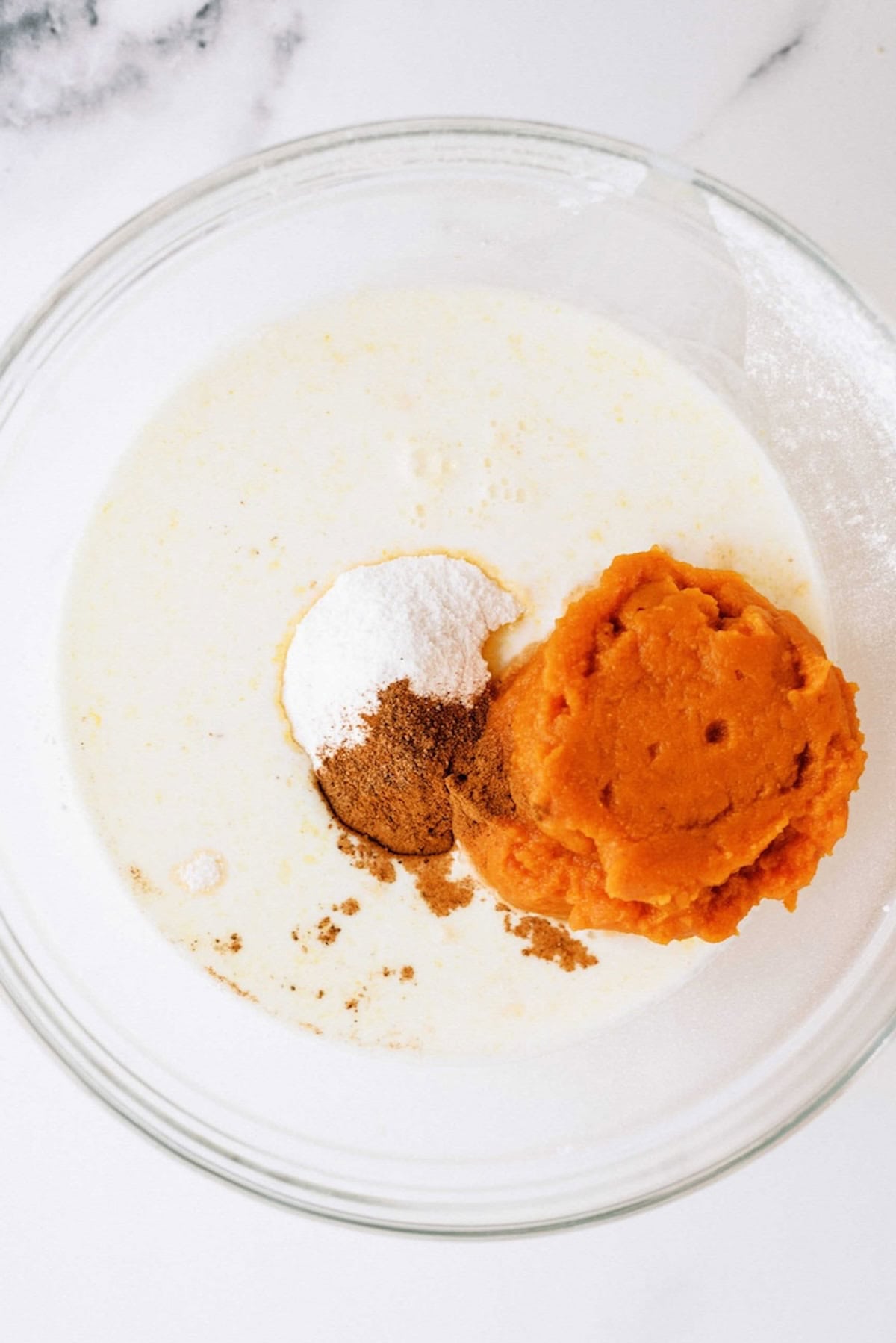 A glass bowl containing milk, a dollop of pumpkin puree, a sprinkle of spices, and a scoop of sugar.