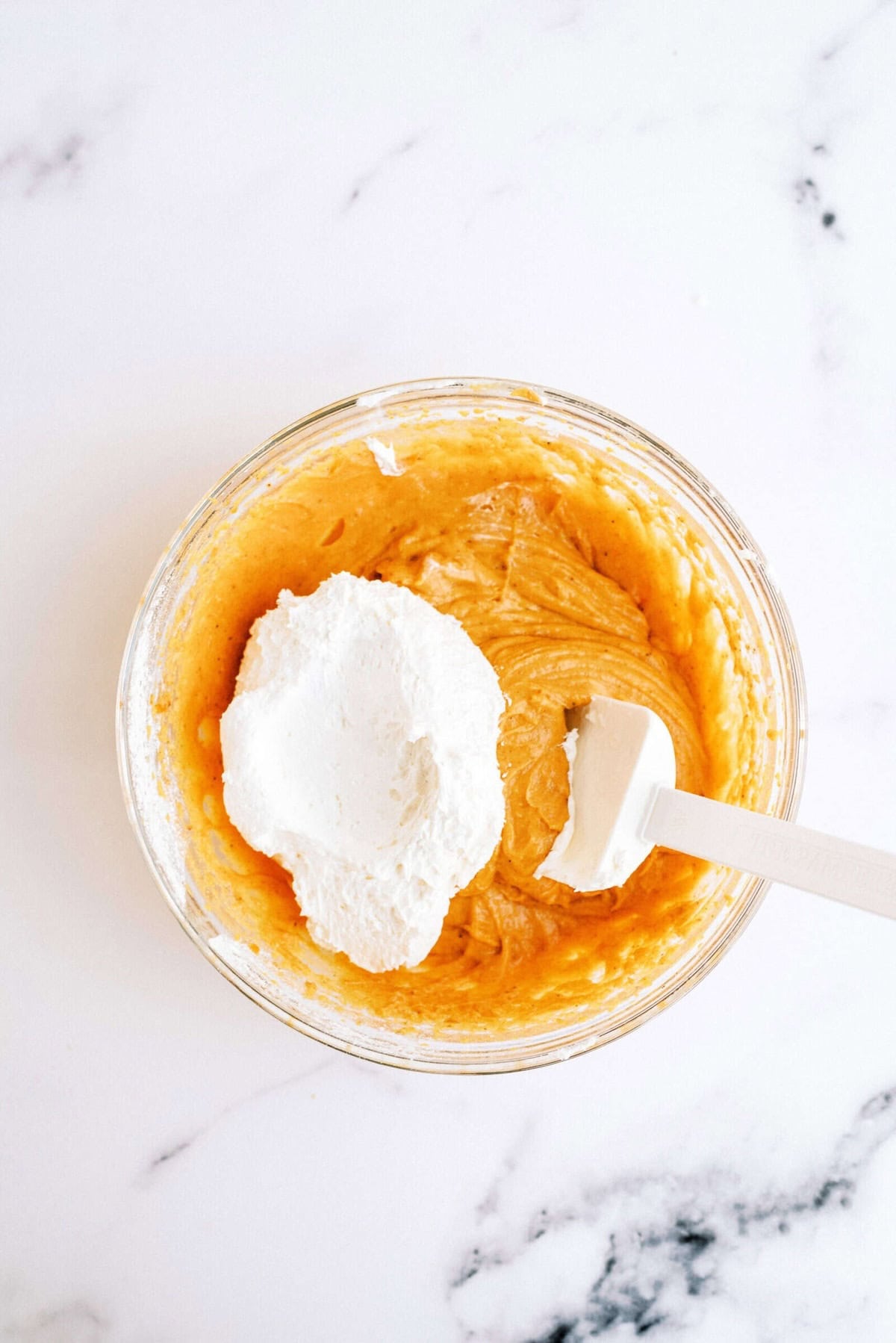 A mixing bowl with an orange batter and a dollop of white cream being folded in with a spatula, sitting on a marble countertop.