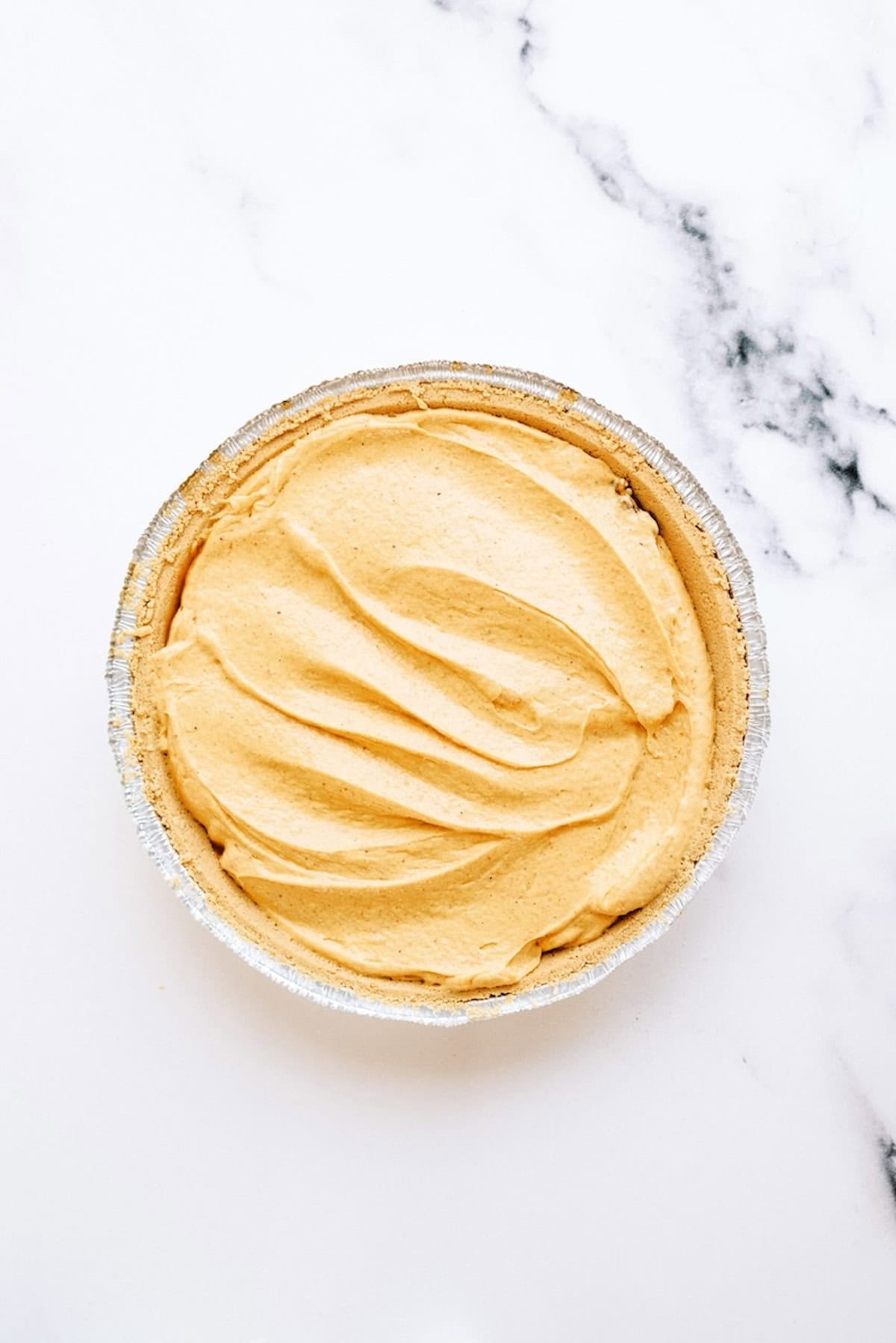 Top-down view of a pumpkin pie with a smooth, creamy filling in a foil pie tin, placed on a white marble surface.