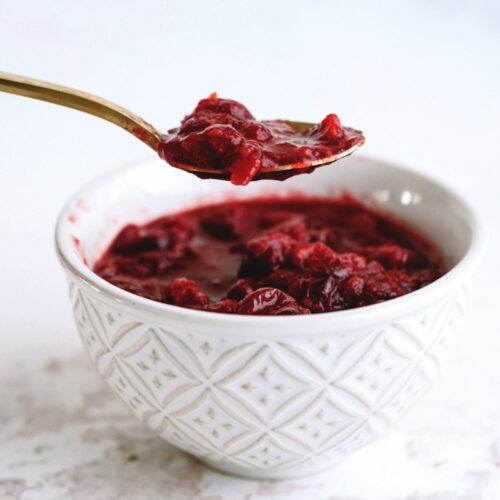 A spoonful of red cranberry sauce held above a white textured bowl filled with cranberry sauce.