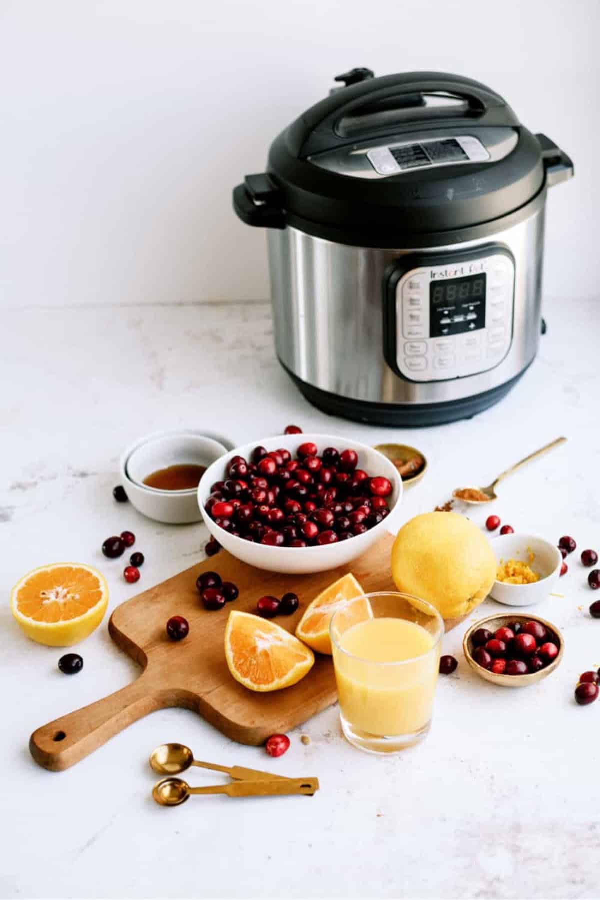 Ingredients including cranberries, oranges, and a lemon on a wooden board with a glass of juice, small bowls, and a pressure cooker in the background.