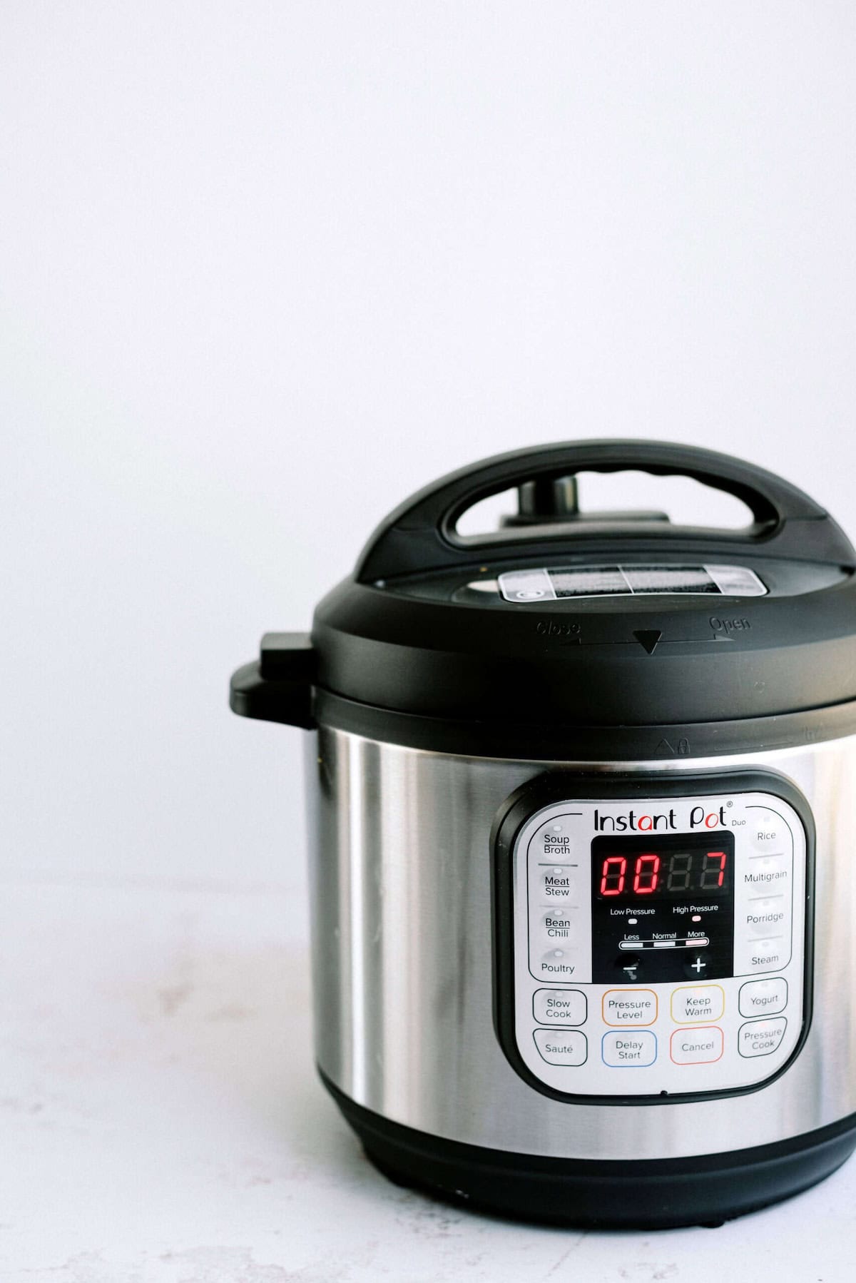 A stainless steel electric pressure cooker sits on a white surface. The display screen shows "00:07," and various cooking function buttons are visible.