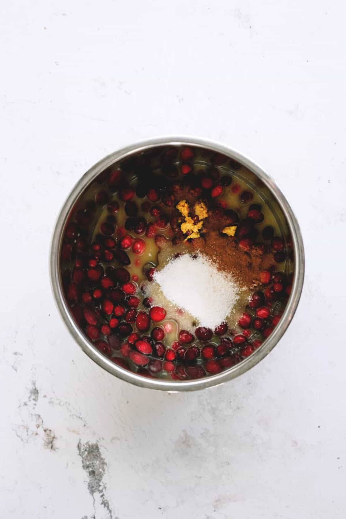 A metal bowl containing cranberries, sugar, spices, and orange zest on a white surface.