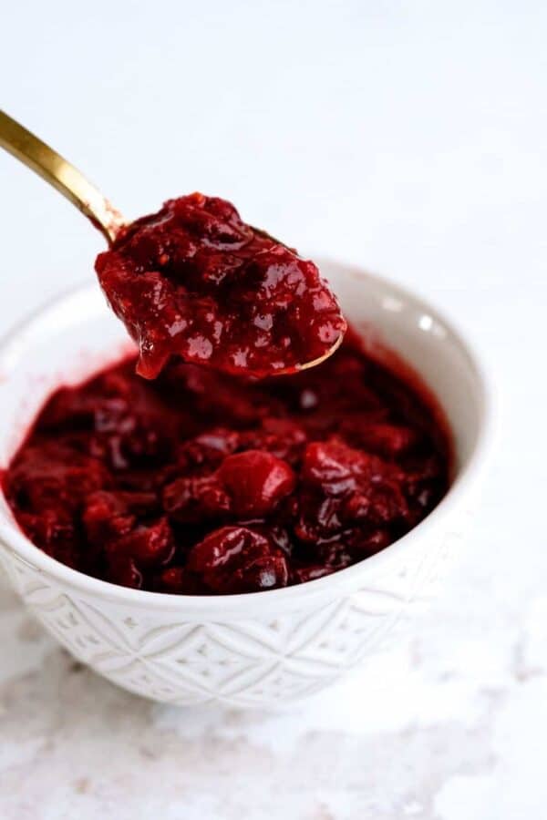 A spoon holds a scoop of red cranberry sauce above a white bowl filled with the same sauce.