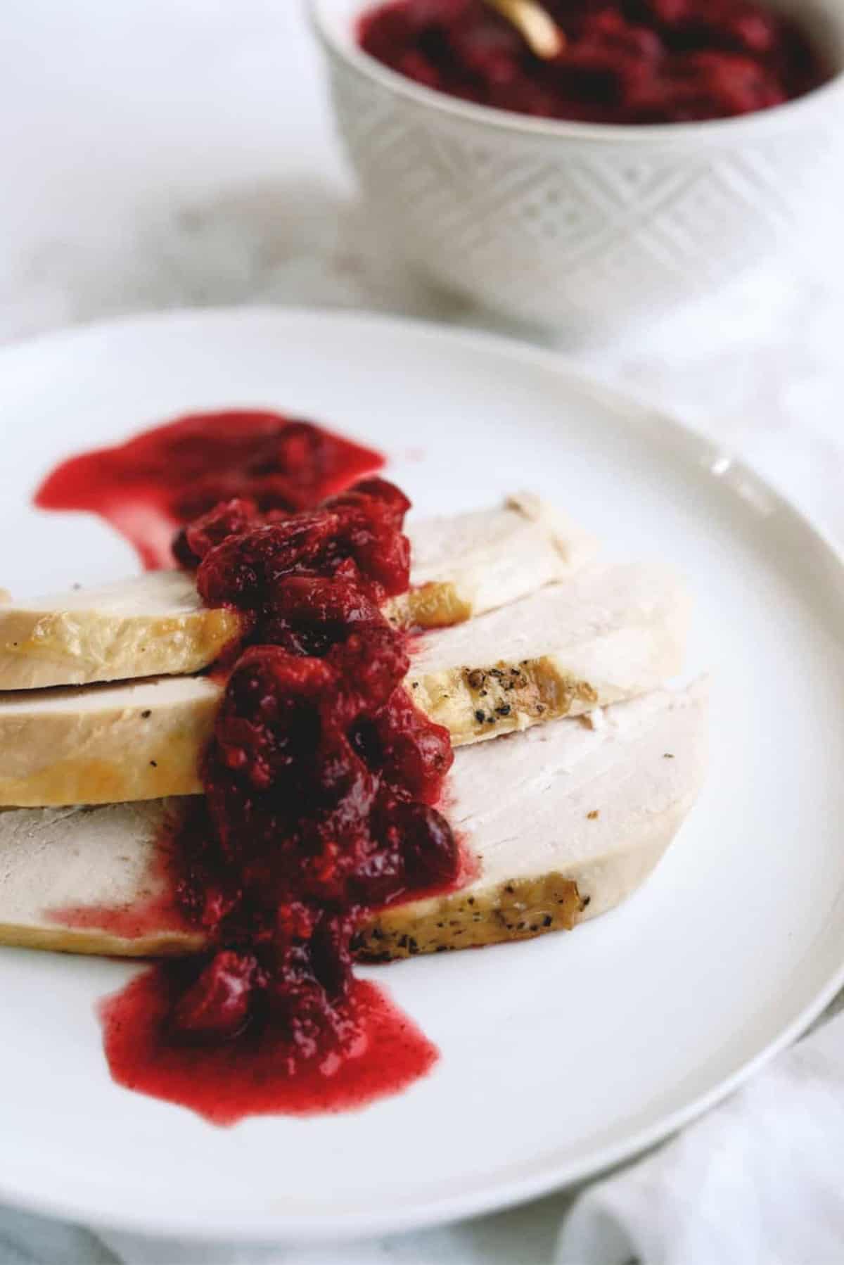 Sliced turkey breast topped with red cranberry sauce on a white plate; a bowl of cranberry sauce in the background.