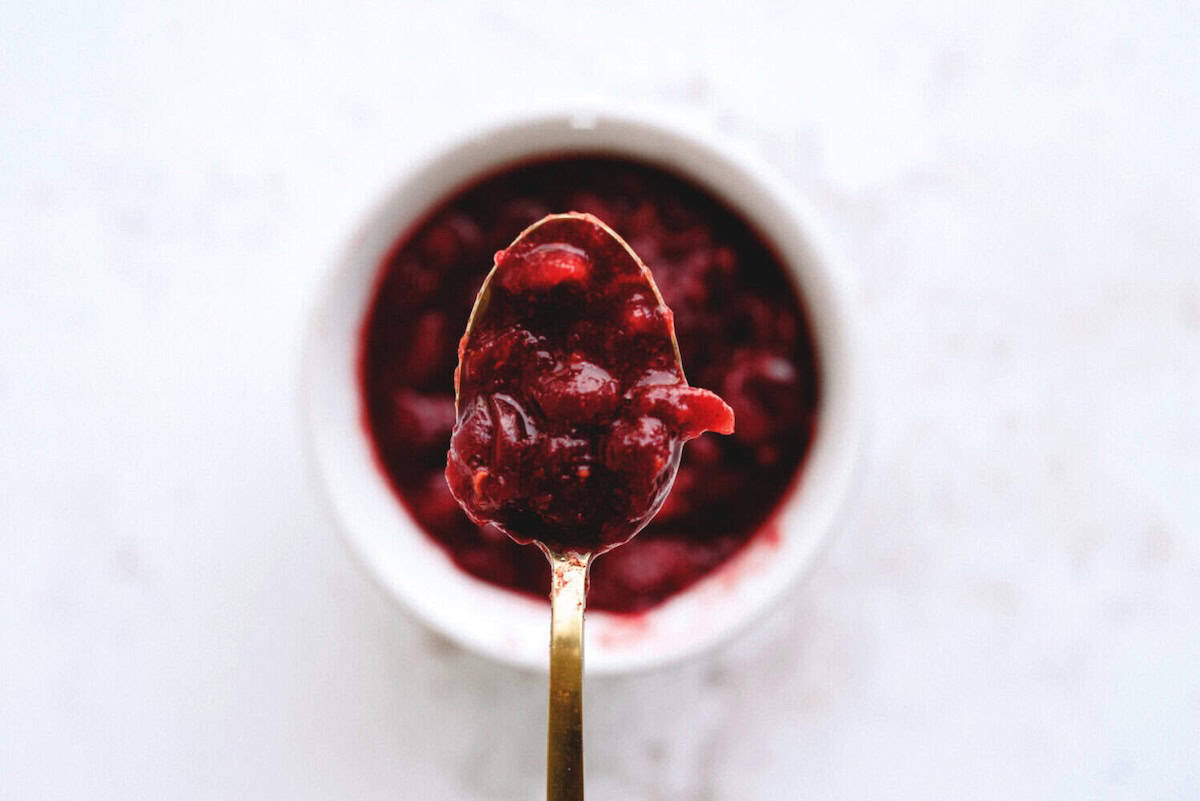 A spoonful of cranberry sauce is held above a white bowl filled with the same jam, on a light background.