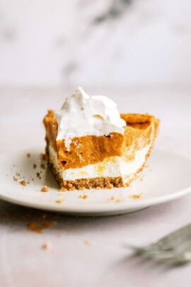 A partially eaten slice of pumpkin pie with whipped cream on top sits on a white plate. Crumbs are scattered around the plate.