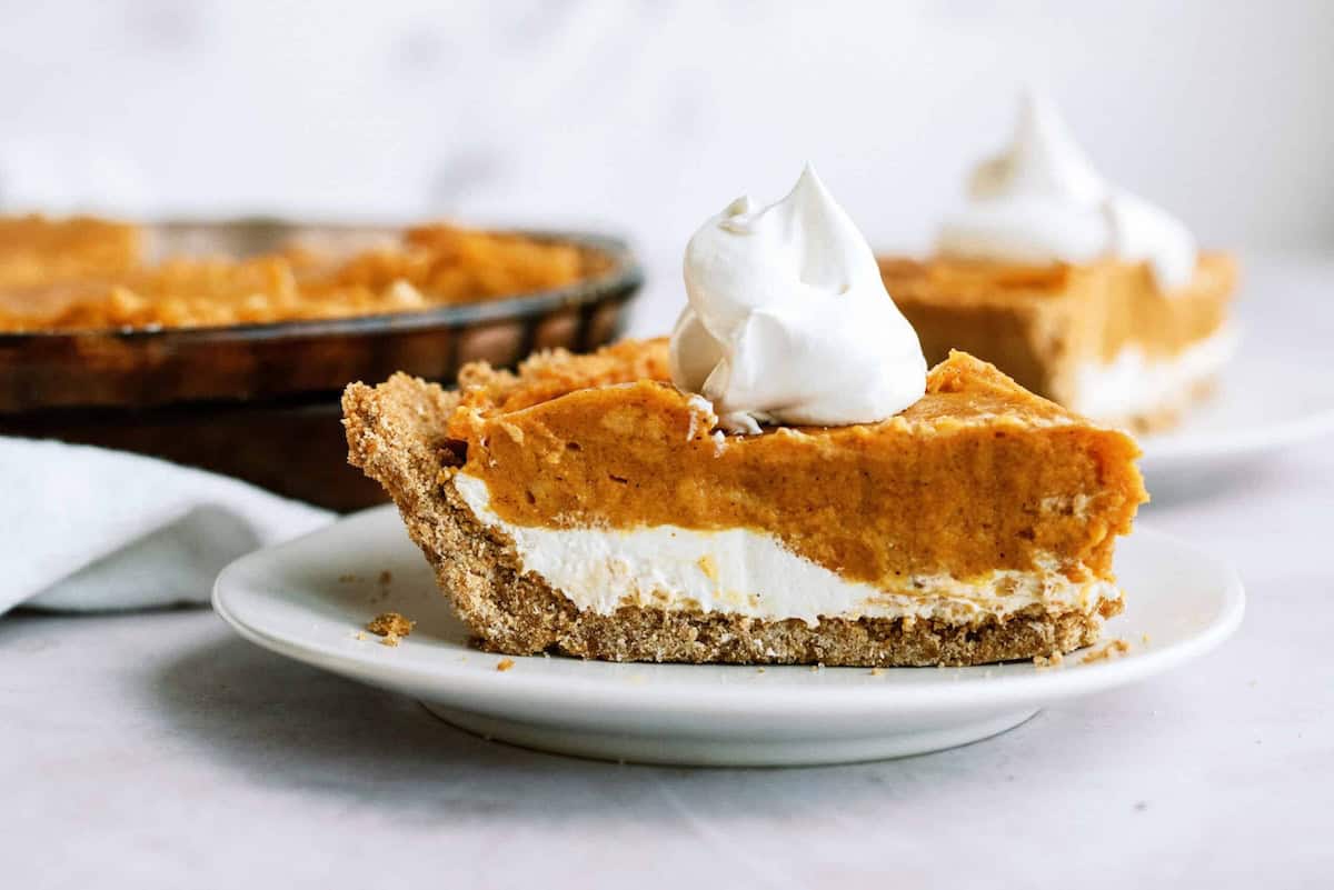 A slice of pumpkin pie with a graham cracker crust and whipped cream topping on a white plate.