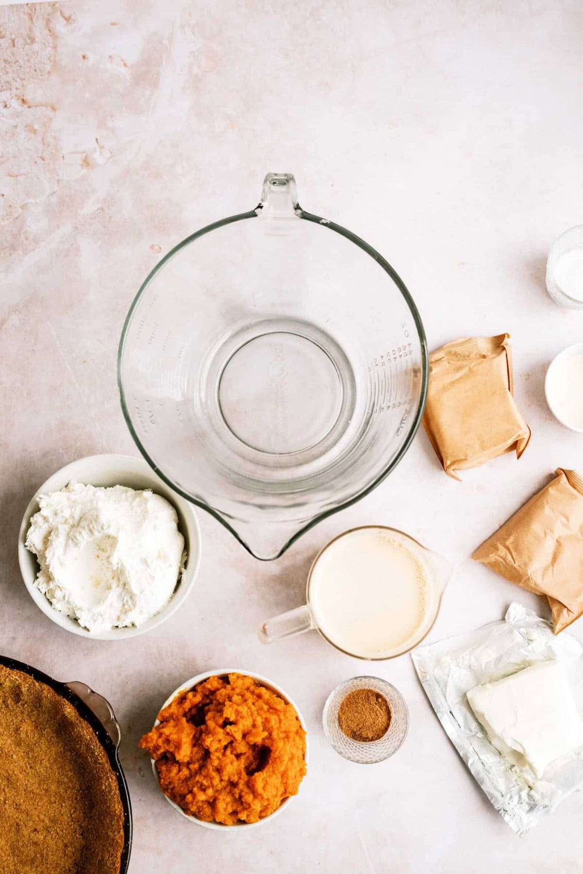 Ingredients for making a pumpkin cheesecake, including a graham cracker crust, bowl of ricotta, pumpkin puree, cream, spices, and packages of cream cheese, arranged around a large glass mixing bowl.