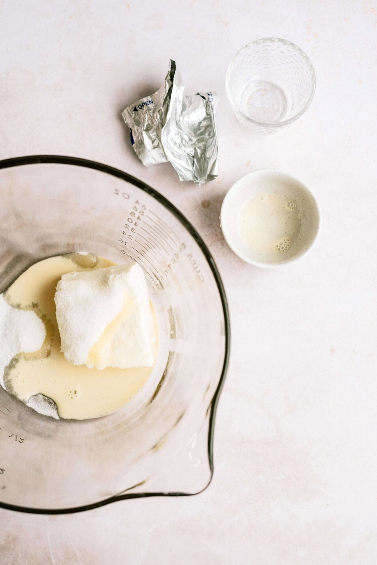 A mixing bowl contains sugar, butter, and a small amount of liquid. Nearby are an empty, unwrapped butter wrapper, a glass, and a small bowl with liquid residue.