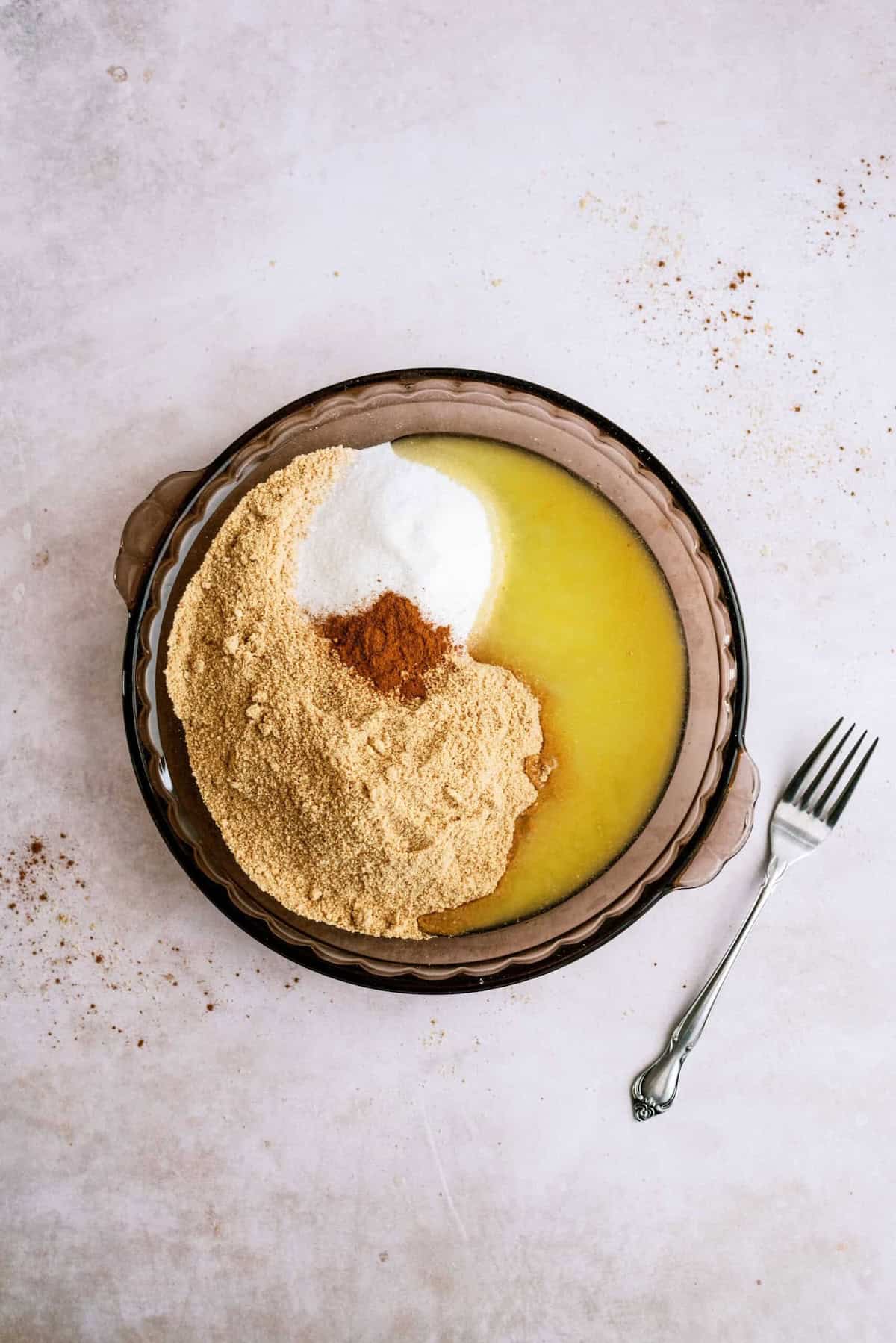 A pie dish containing melted butter, graham cracker crumbs, sugar, and cinnamon, with a fork placed on the right side.