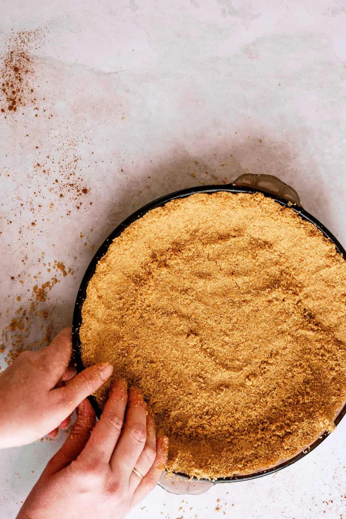 Hands pressing a graham cracker crust into a round pie dish on a light-colored surface with some crumbs scattered around.