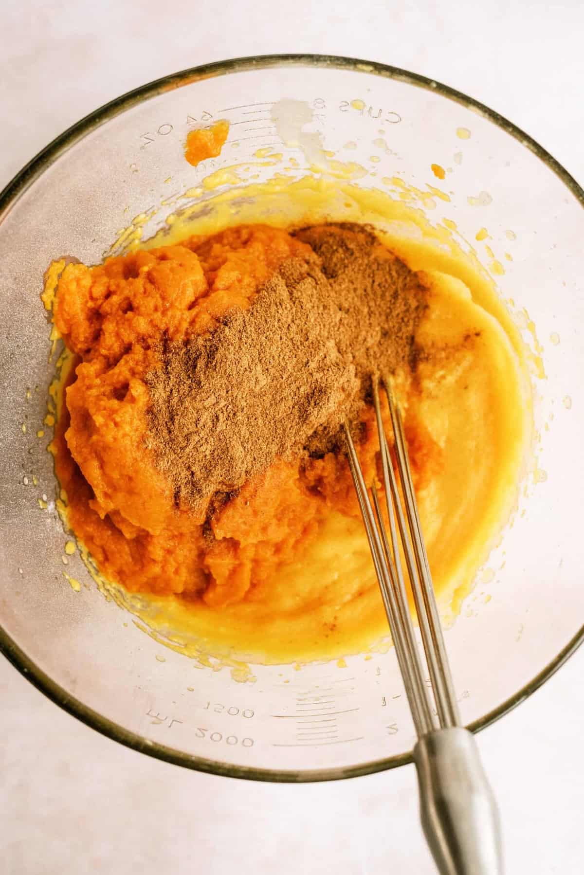 A mixing bowl with pumpkin puree, brown sugar, and a whisk partially submerged in the mixture, on a light-colored background.