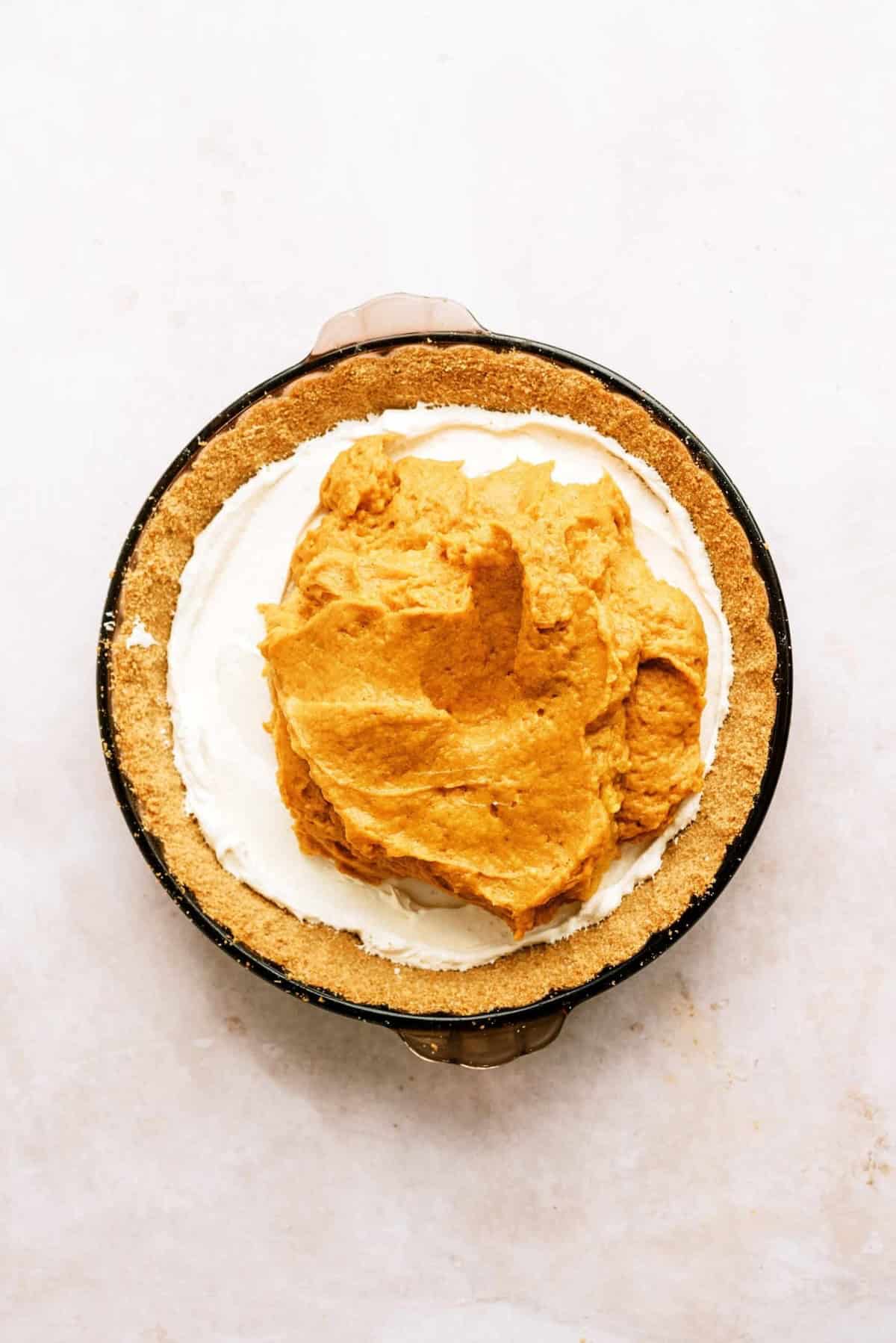 A pie dish with a graham cracker crust is filled with a layer of white and a dollop of orange filling yet to be spread.