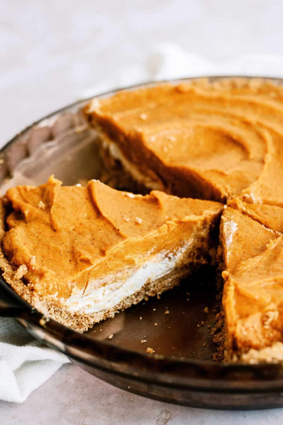 A clear pie dish containing a pumpkin pie with a swirl pattern and a graham cracker crust, showing a few slices removed.