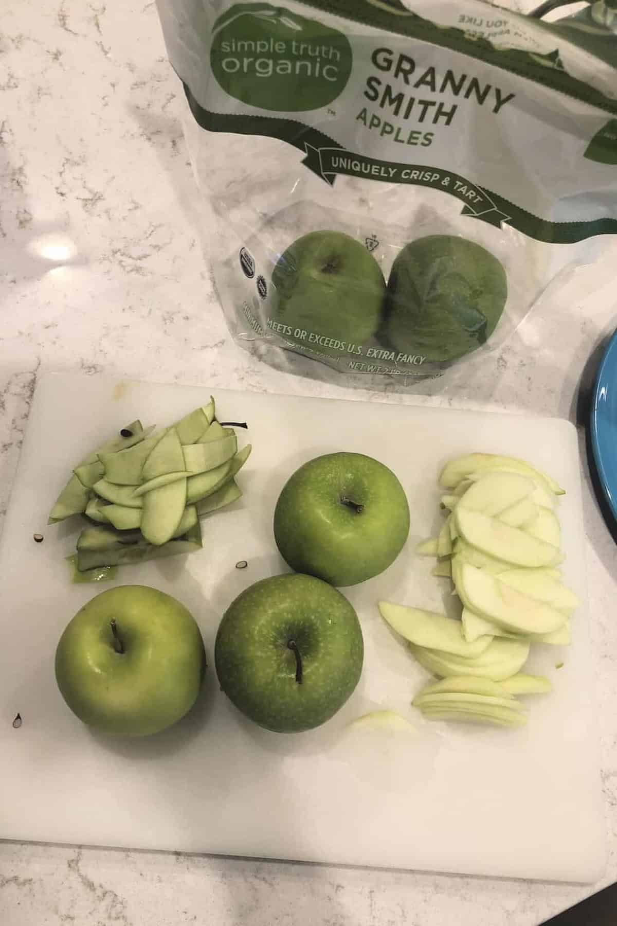 Sliced and whole Granny Smith apples on a white cutting board, with a package labeled "Simple Truth Organic Granny Smith Apples" in the background.