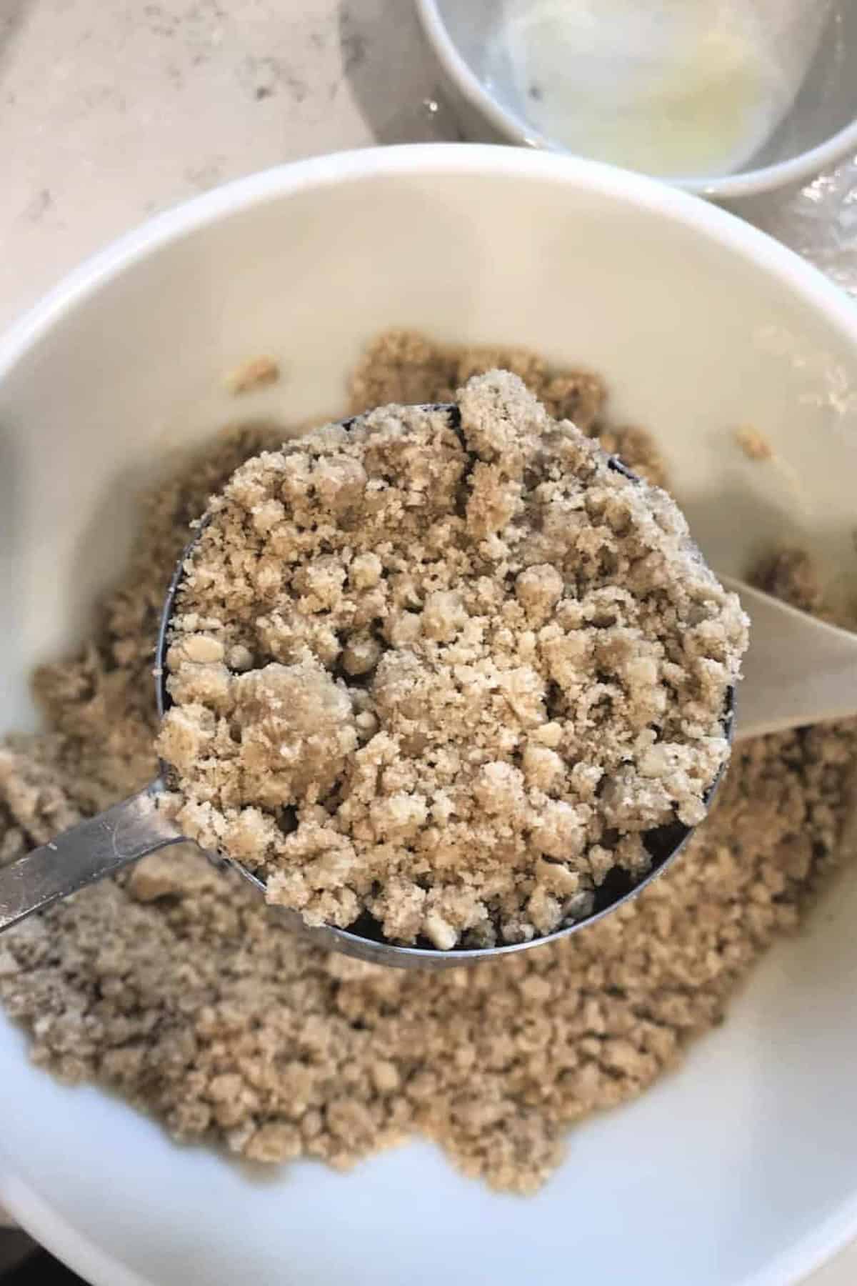 A measuring cup filled with crumbly mixture is above a white bowl containing more of the mixture.