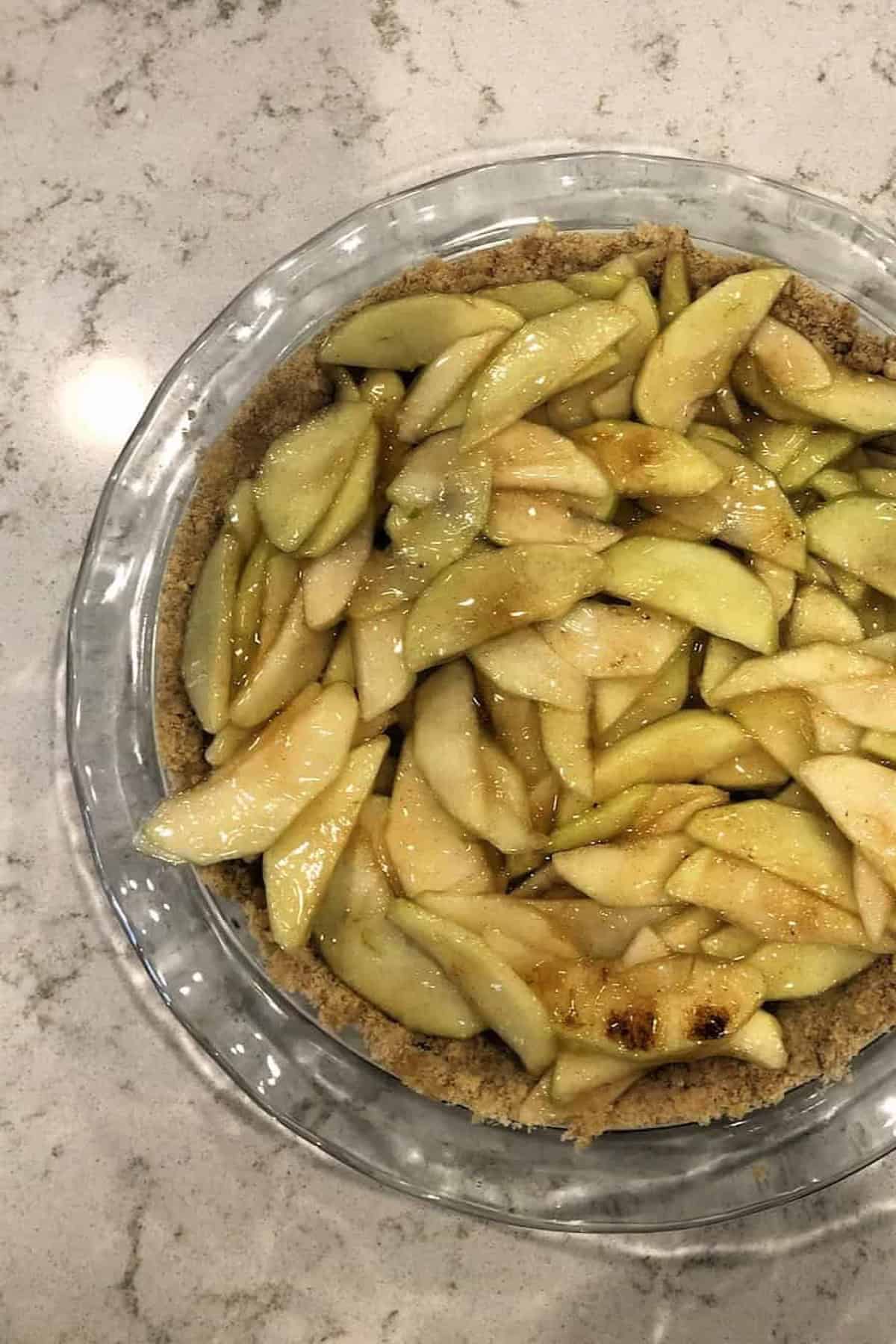 Pie with sliced apples on a crust in a glass dish, placed on a marble countertop.