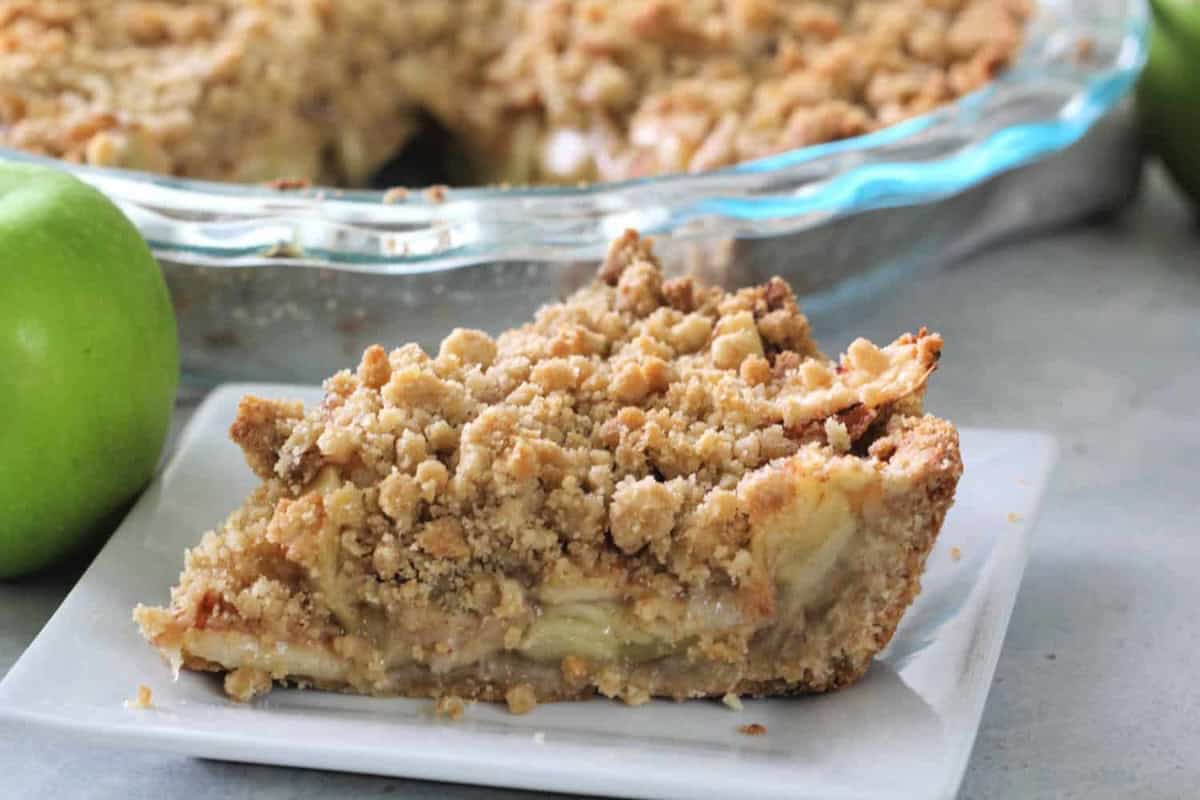 A slice of apple crumble pie on a small white plate, with the rest of the pie in a glass dish in the background. Green apples are partially visible.