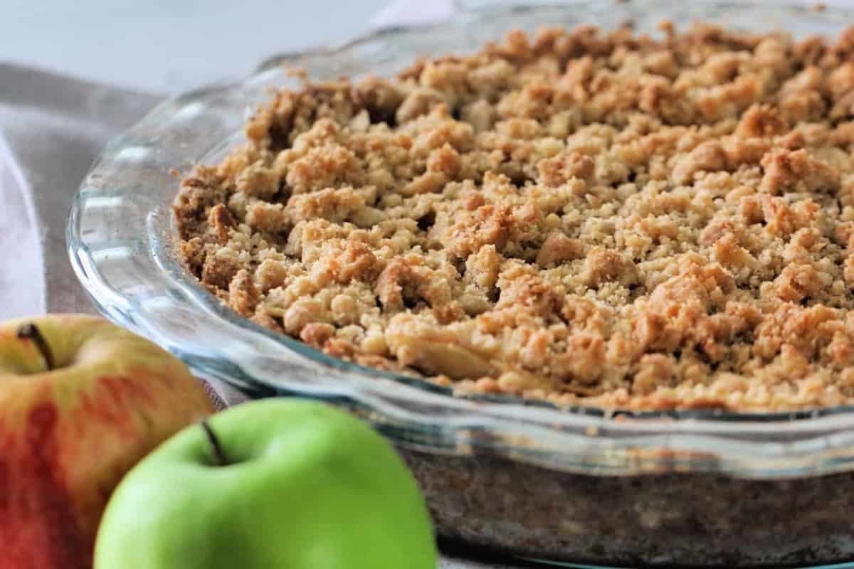 A glass dish contains a baked apple crumble with a golden, crumbly top. Two apples, one red and one green, are placed beside the dish on a light surface.