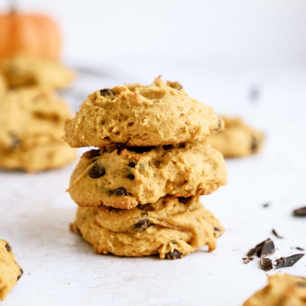 A stack of three pumpkin chocolate chip cookies on a white surface with more cookies and chocolate chips scattered around in the background.