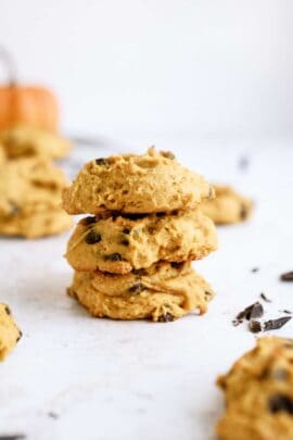 A stack of three pumpkin chocolate chip cookies on a white surface with more cookies and chocolate chips scattered around in the background.