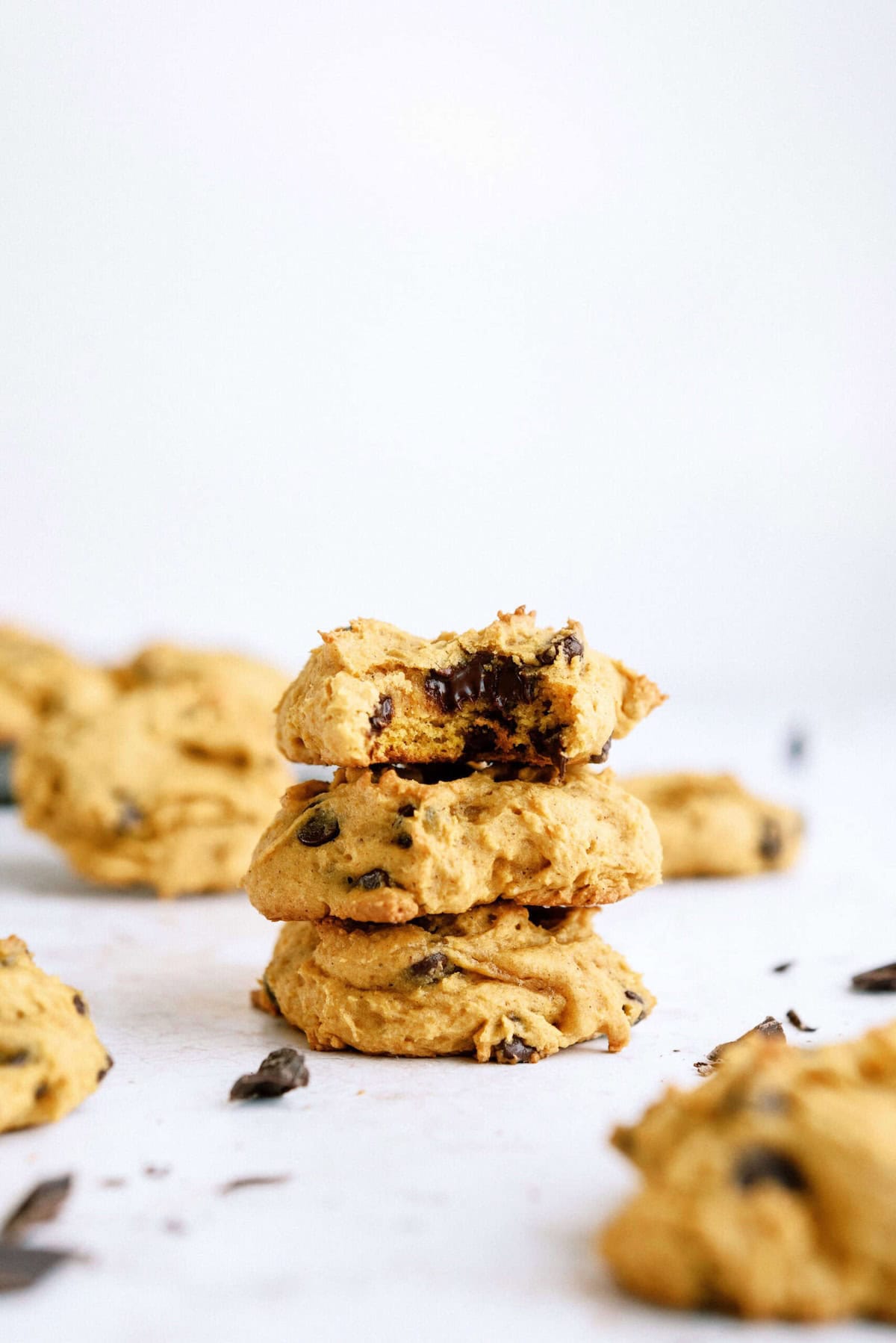 A stack of three pumpkin chocolate chip cookies, the top one with a bite taken out, surrounded by more cookies on a white surface.