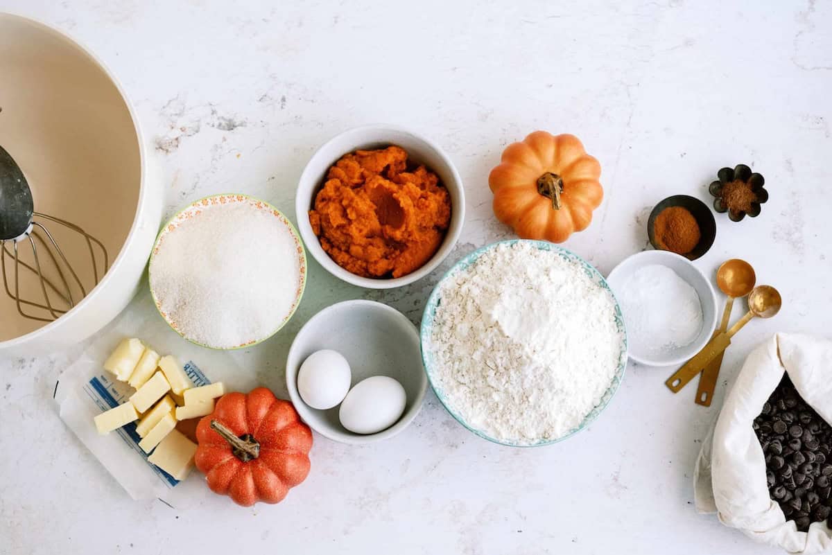 Various baking ingredients are spread out on a white surface, including flour, sugar, eggs, butter, pumpkin puree, and chocolate chips. There are also measuring spoons and autumn-themed items.