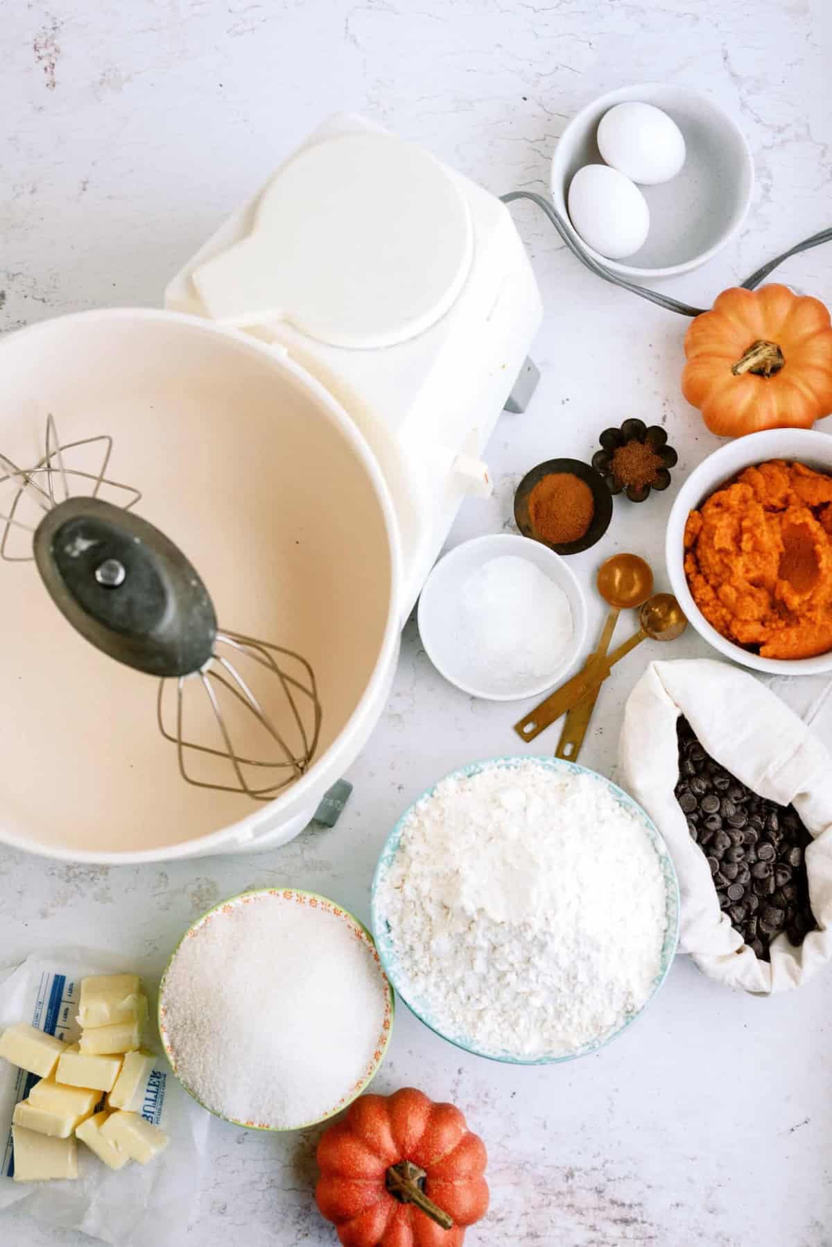 A stand mixer with a bowl is surrounded by ingredients for baking, including flour, butter, sugar, chocolate chips, pumpkin puree, eggs, spices, and measuring spoons arranged on a white countertop.