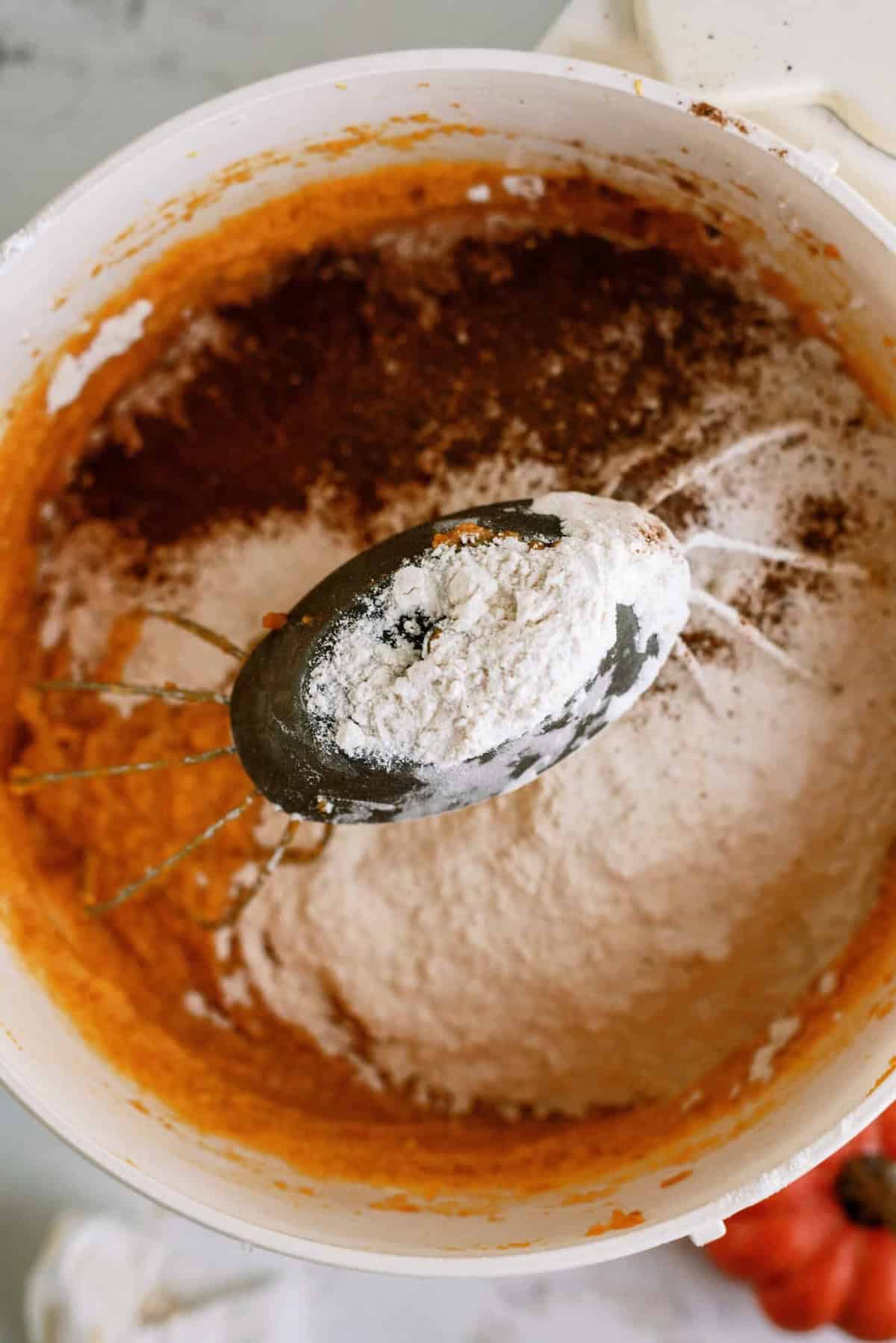 A mixing bowl containing pumpkin batter with flour and spices, partially mixed, with beaters in the bowl.