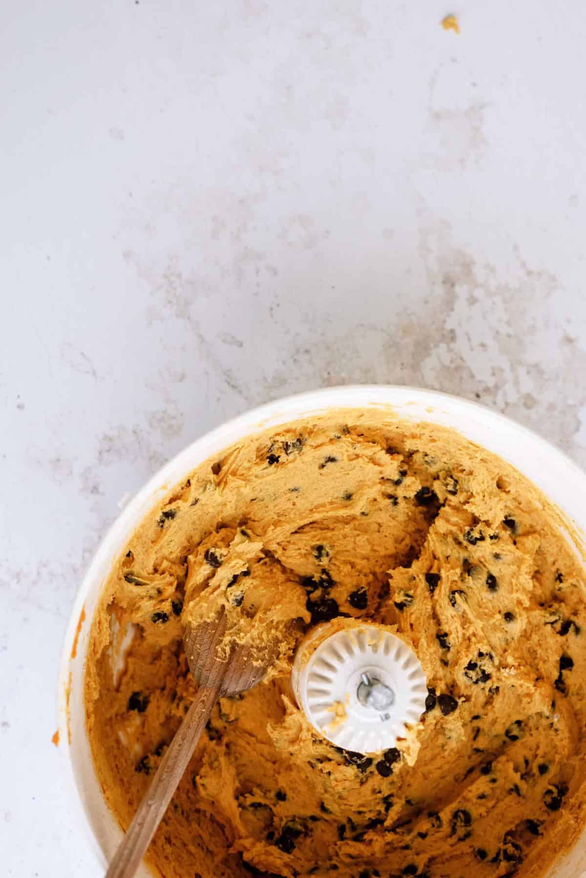 A bowl of cookie dough with chocolate chips and a wooden spoon inside, resting on a white surface.