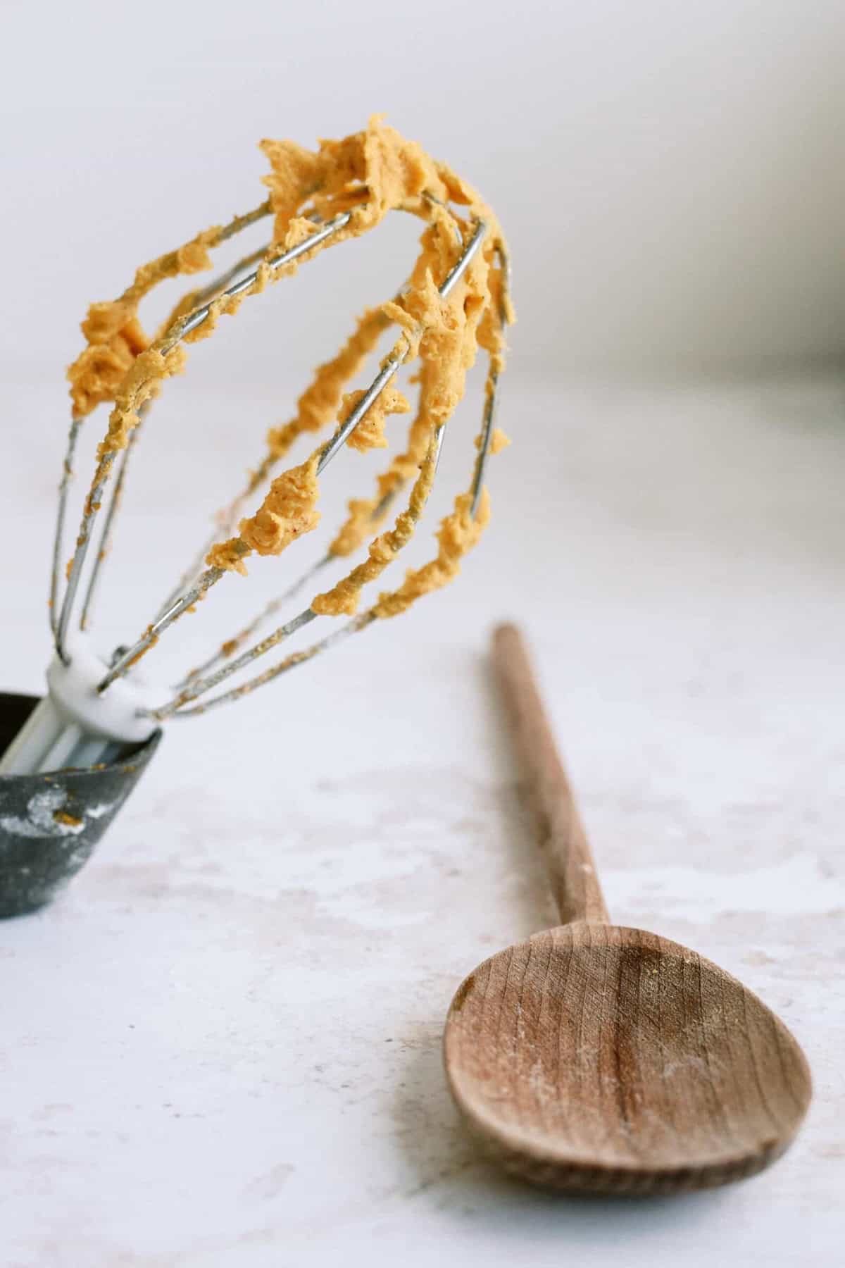 A whisk covered in dough rests on a countertop next to a wooden spoon.