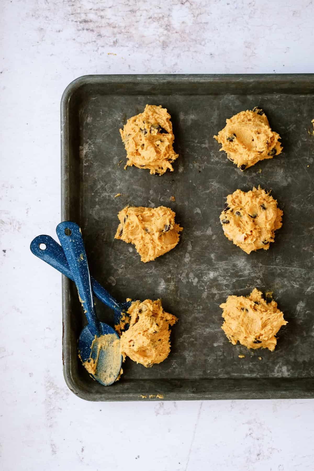 Six dollops of cookie dough arranged on a dark baking sheet, with two blue spoons covered in dough resting on the sheet.