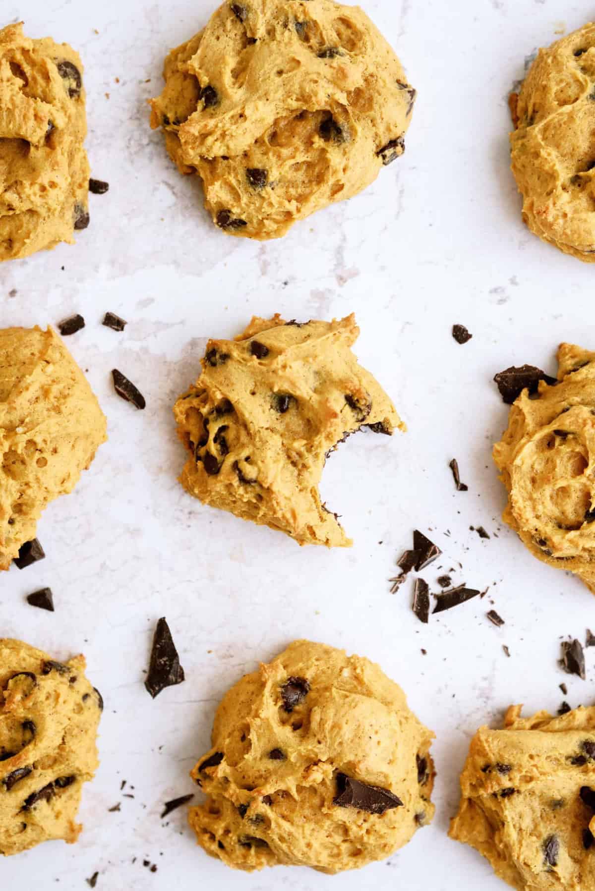 Nine pumpkin chocolate chip cookies and some chocolate chunks are spread on a white surface. One cookie has a bite taken out of it.