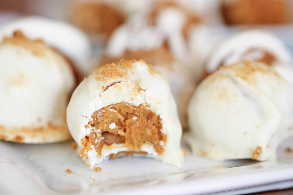 Close-up of several round white chocolate-covered truffles, with one partially bitten to reveal a brown crumbly filling. The cookies are displayed on a white surface.