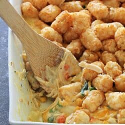 A close-up of a casserole dish filled with tater tots, vegetables, and creamy sauce, with a wooden spoon scooping a portion.